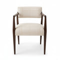 A wooden armchair with cream-colored textured fabric upholstery, shown in a home setting on a wood floor near a window.